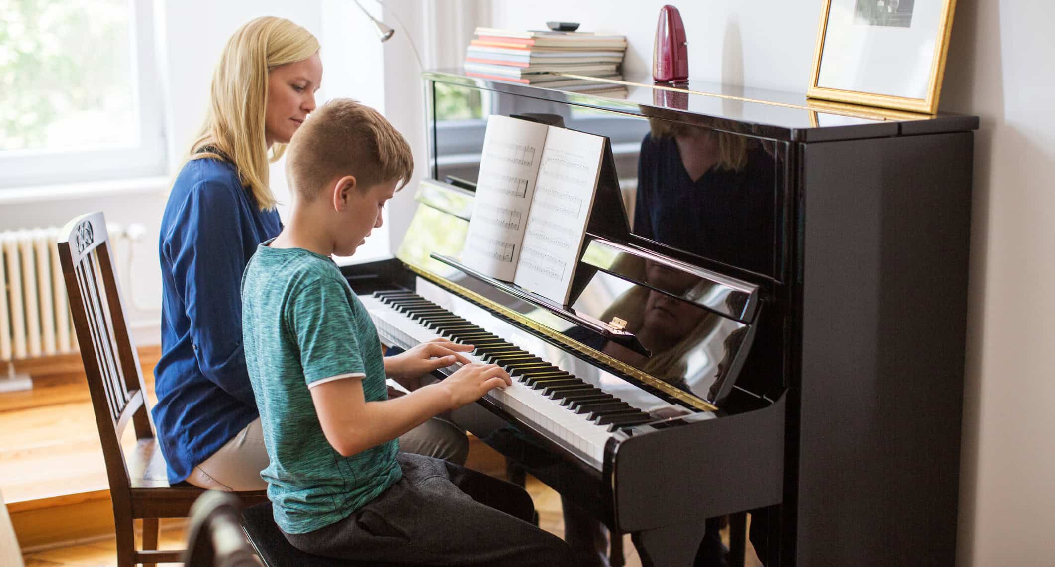 Young boy learning to play beginner piano on upright as mom looks on